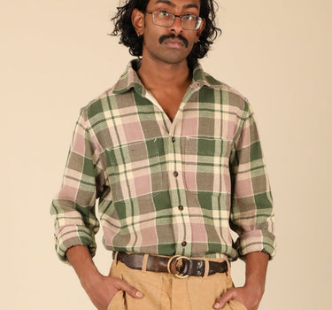A man modeling a green and pink plaid button-down shirt with tan pants, brown shoes, and a brown belt, standing against a solid beige background.