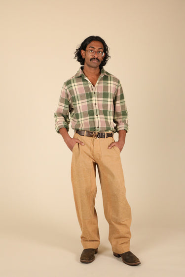 A man modeling a green and pink plaid button-down shirt with tan pants, brown shoes, and a brown belt, standing against a solid beige background.
