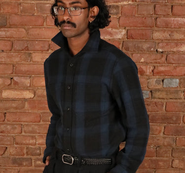 A man wearing a dark blue and black plaid button-down shirt, black trousers, and black dress shoes stands in front of a red brick wall and wooden floor.