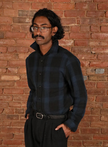 A man wearing a dark blue and black plaid button-down shirt, black trousers, and black dress shoes stands in front of a red brick wall and wooden floor.