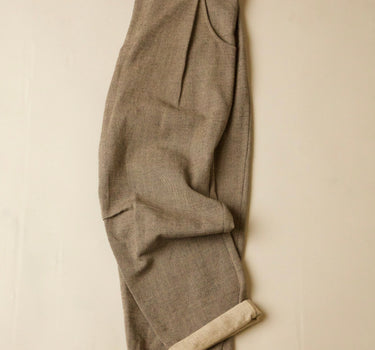 A pair of grey pleated trousers with a rolled-up hem showing a lighter beige lining, laid flat against a neutral cream background.