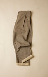 A pair of grey pleated trousers with a rolled-up hem showing a lighter beige lining, laid flat against a neutral cream background.