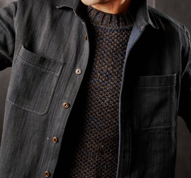 A person wearing an iron/indigo denim workshirt with chest pockets and Mother of Pearl buttons, paired with a knit sweater.