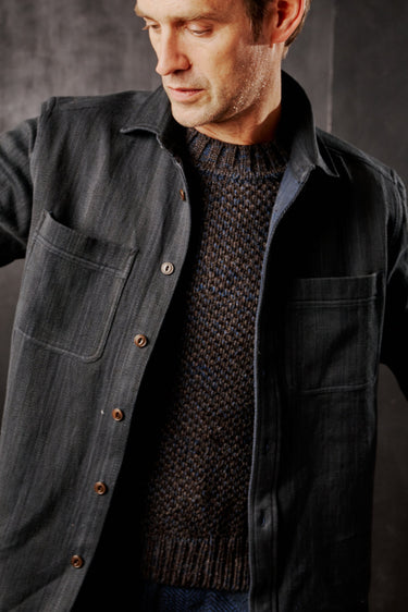 A person wearing an iron/indigo denim workshirt with chest pockets and Mother of Pearl buttons, paired with a knit sweater.