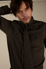 A man wearing a dark brown pinstriped button-down shirt and matching tie, posing with one arm behind his head against a plain beige background.