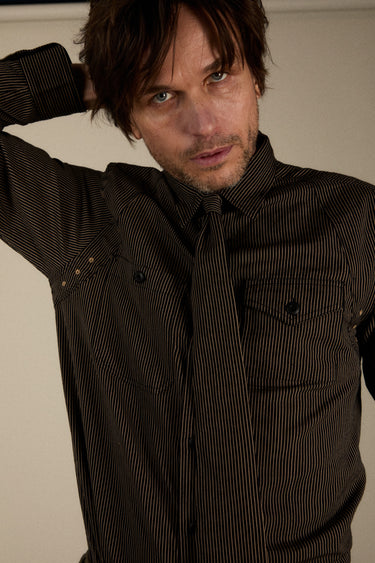 A man wearing a dark brown pinstriped button-down shirt and matching tie, posing with one arm behind his head against a plain beige background.