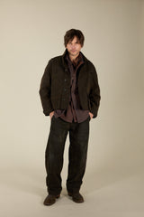 A man modeling a dark brown utility jacket over a dark brown button-down shirt and dark charcoal pants, standing against a plain beige studio background.