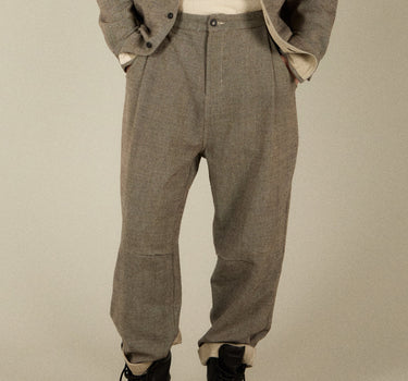 A man modeling a matching two-piece outfit consisting of a gray tweed button-up overshirt and matching gray trousers, styled with a cream crewneck t-shirt and black boots, against a plain beige studio background.