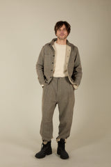 A man modeling a matching two-piece outfit consisting of a gray tweed button-up overshirt and matching gray trousers, styled with a cream crewneck t-shirt and black boots, against a plain beige studio background.