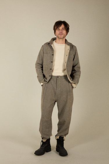 A man modeling a matching two-piece outfit consisting of a gray tweed button-up overshirt and matching gray trousers, styled with a cream crewneck t-shirt and black boots, against a plain beige studio background.