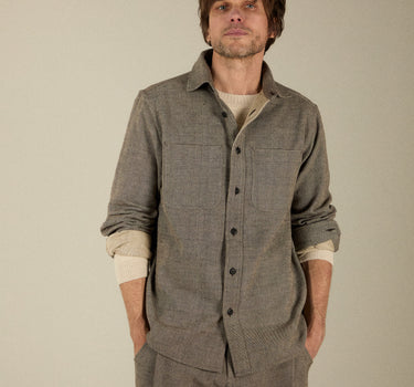 A man wearing a textured gray button-up shirt and matching gray trousers over a cream undershirt, standing against a solid light beige background.