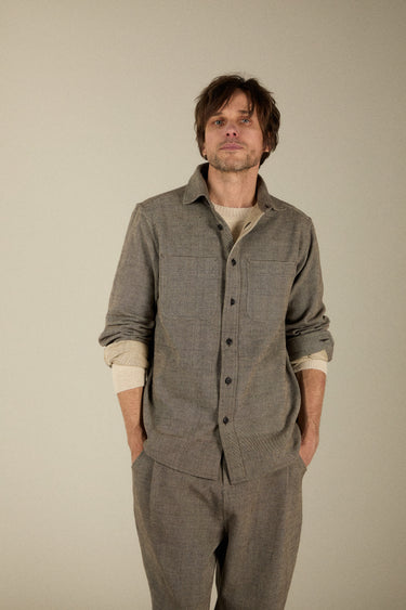 A man wearing a textured gray button-up shirt and matching gray trousers over a cream undershirt, standing against a solid light beige background.