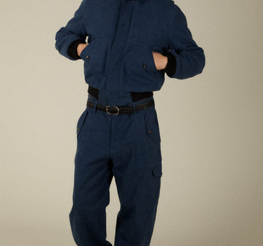 A man modeling a dark navy blue heavy-duty workwear set consisting of a cropped jacket with black ribbed collar and cuffs, and matching dark blue trousers, against a plain beige studio background.
