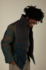 A man wearing a textured, multi-colored patchwork jacket with blue, green, and brown tones, styled with a dark shirt and beige trousers against a plain beige background.