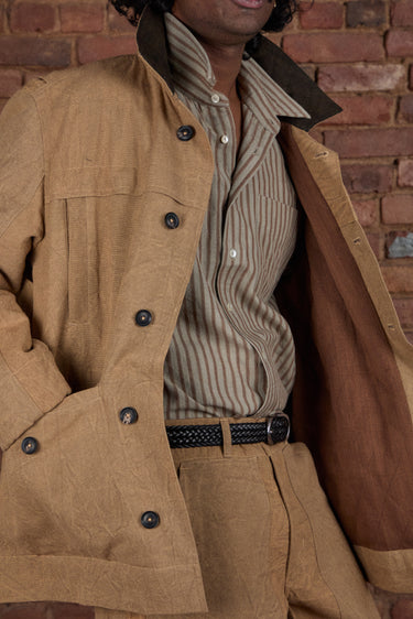 A close-up of a person wearing a tan canvas utility jacket with dark buttons and a brown collar, layered over a beige and brown vertically striped button-down shirt, set against a rustic red brick wall.