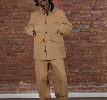 A man wearing a tan two-piece linen suit with a band-collar jacket and matching trousers, standing in front of a red brick wall and wooden floor.