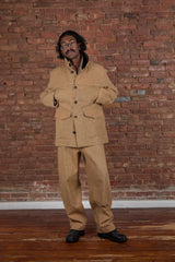 A man wearing a tan two-piece linen suit with a band-collar jacket and matching trousers, standing in front of a red brick wall and wooden floor.