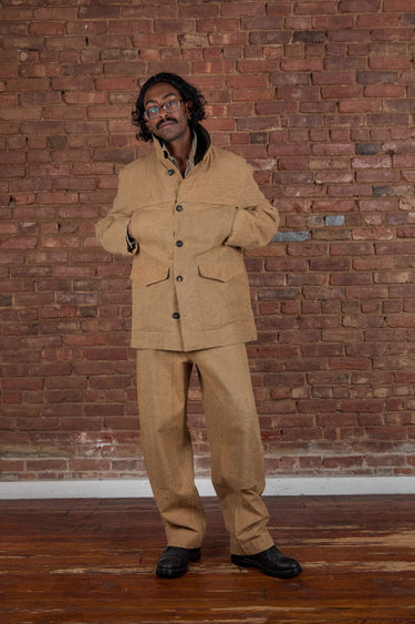 A man wearing a tan two-piece linen suit with a band-collar jacket and matching trousers, standing in front of a red brick wall and wooden floor.