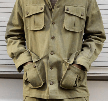 A man wearing a muted olive green military-style jacket with four front pockets and matching pants, standing against a white paneled wall.