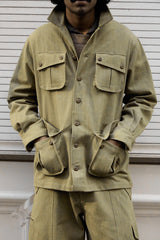 A man wearing a muted olive green military-style jacket with four front pockets and matching pants, standing against a white paneled wall.