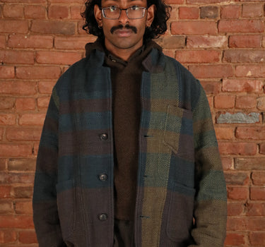 A man wearing a plaid overshirt jacket in muted blue, green, and brown tones, layered over a dark brown sweater and black pants, standing against a red brick wall.