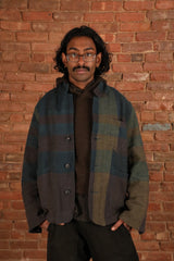 A man wearing a plaid overshirt jacket in muted blue, green, and brown tones, layered over a dark brown sweater and black pants, standing against a red brick wall.